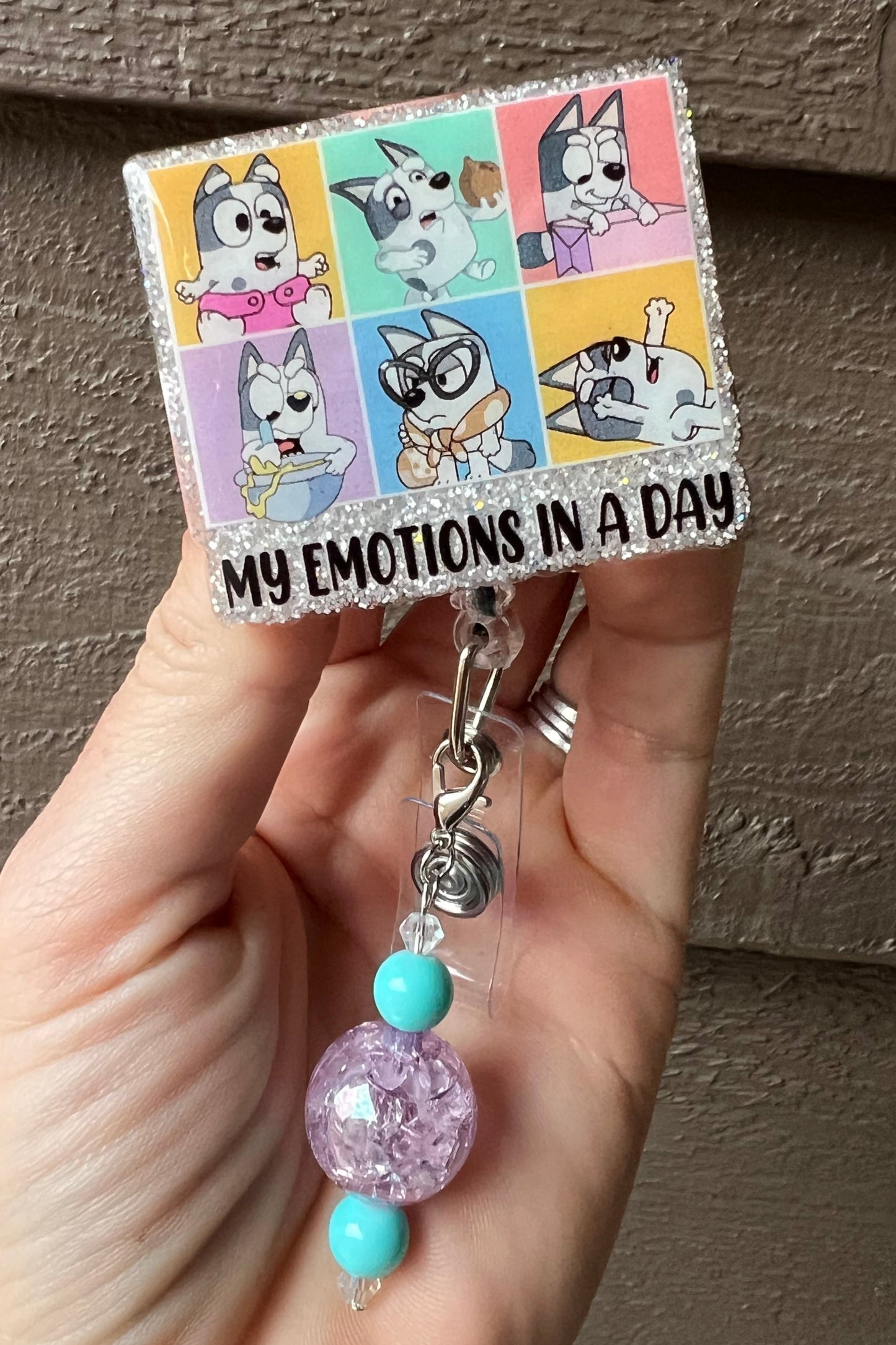 Muffin from Bluey glitter badge reel showing six emotion faces with "My emotions in a day" text and purple beaded dangle.
