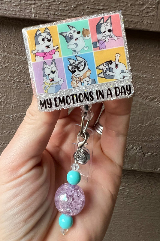 Muffin from Bluey glitter badge reel showing six emotion faces with "My emotions in a day" text and purple beaded dangle.