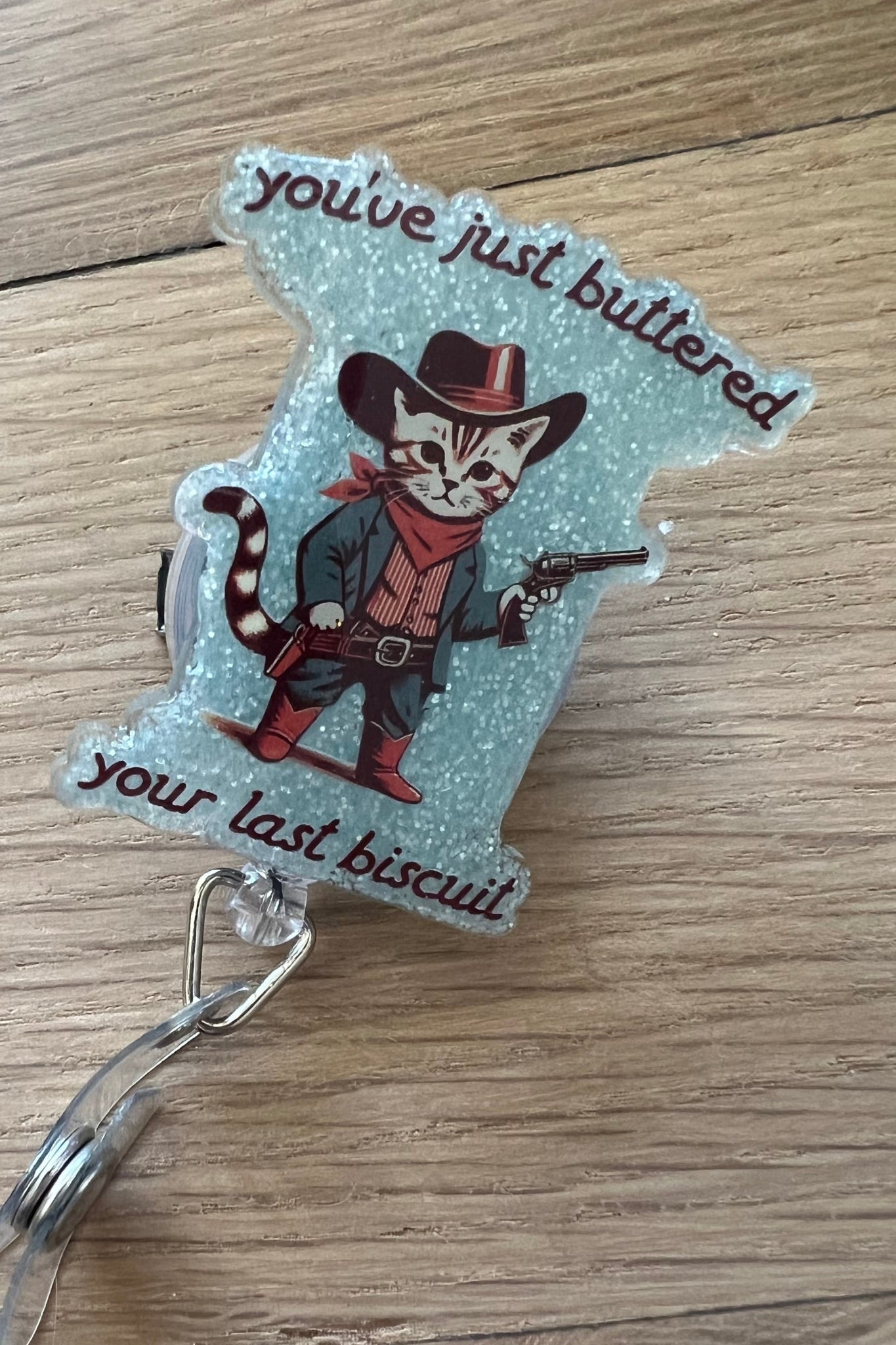 Funny badge reel with cowboy cat and the phrase “You’ve just buttered your last biscuit” on a glittery background