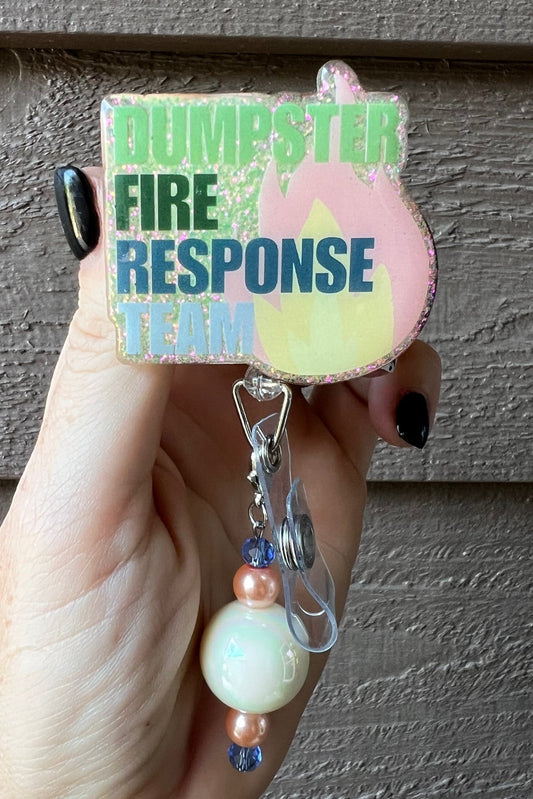 Hand holding soft yellow badge reel with the words "Dumpster Fire Response Team" and soft flame on the right side with coordinating badge bling.