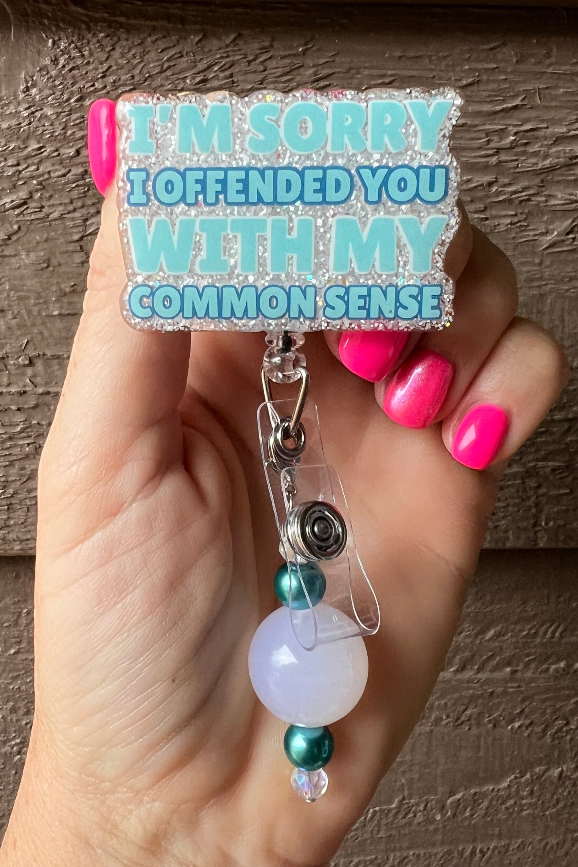 Funny Badge Reel with the phrase "I'm Sorry I Offended You With My Common Sense" in teal/mint text on soft white glitter with white and teal beaded dangle.