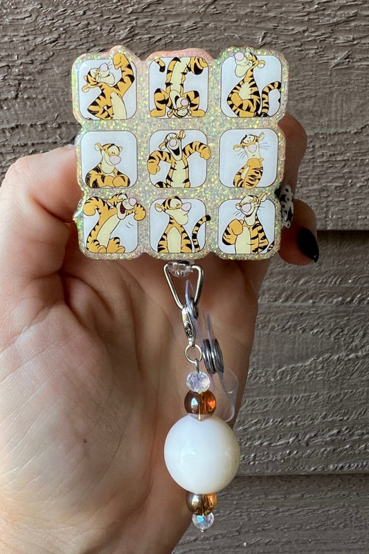 Hand holding Tigger Badge reel with 9 different Tigger poses on soft orange glitter with coordinating badge bling.