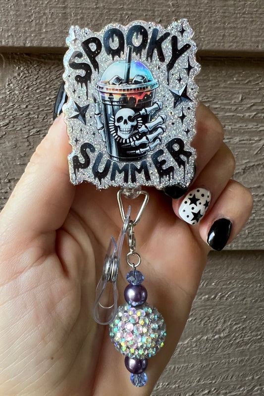 Hand holding badge reel with holographic background and the text "Spooky Summer" with skeleton hand holding an iced drink.