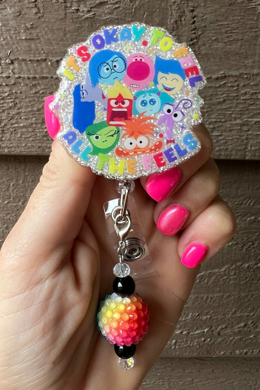 White glitter badge reel featuring the cartoon characters from Inside Out 2, with the text "It's okay to feel all the feels."