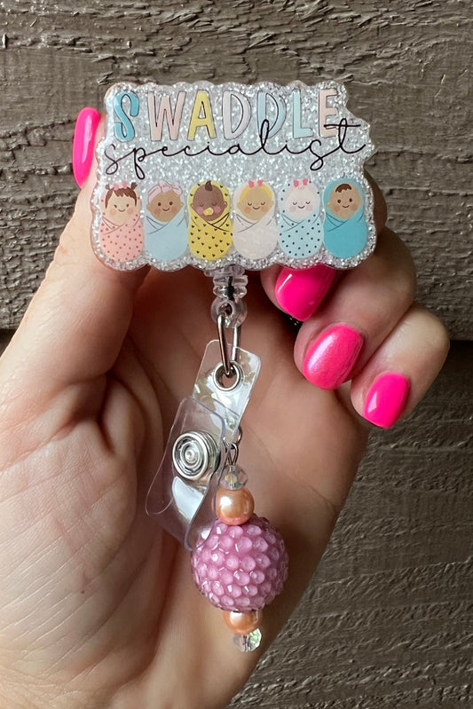 Soft Glitter Badge Reel that says "Swaddle Specialist" with illustrations of six swaddled babies with pink and gold beaded dangle.