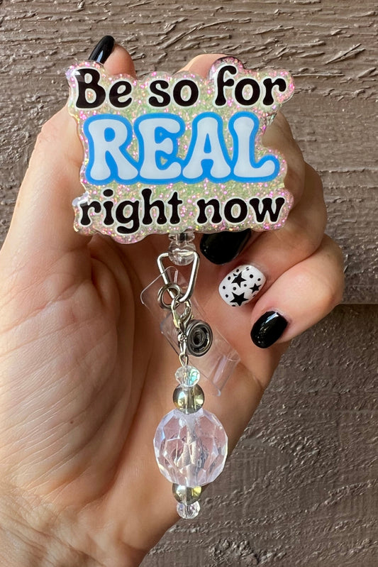 Hand holding soft yellow badge reel with the words "Be so for REAL right now" with coordinating badge bling.