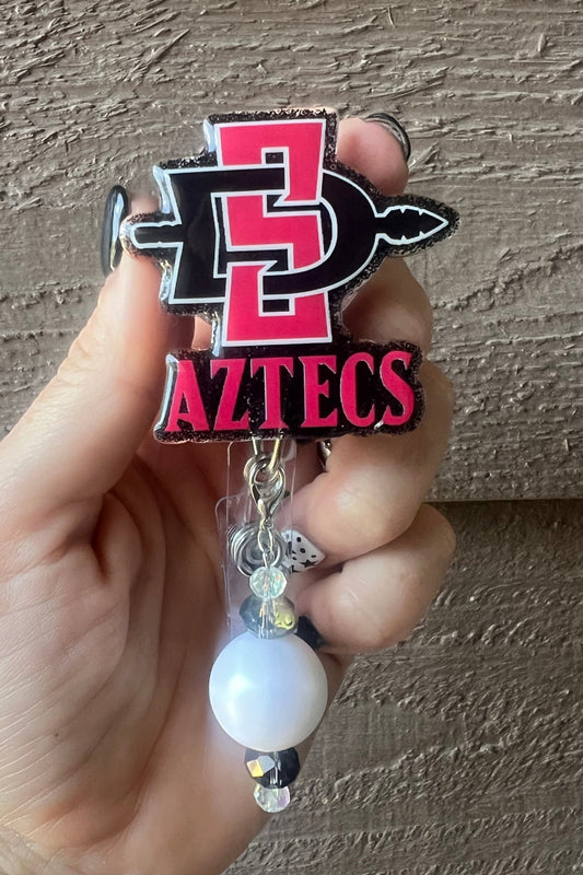 Hand holding badge reel featuring the San Diego State Aztecs logo on black glitter with coordinating badge bling.