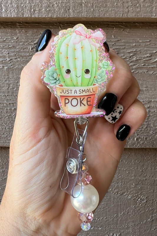 Hand holding cactus badge reel with the phrase "Just a small poke" on the pot of the cactus with matching badge bling.