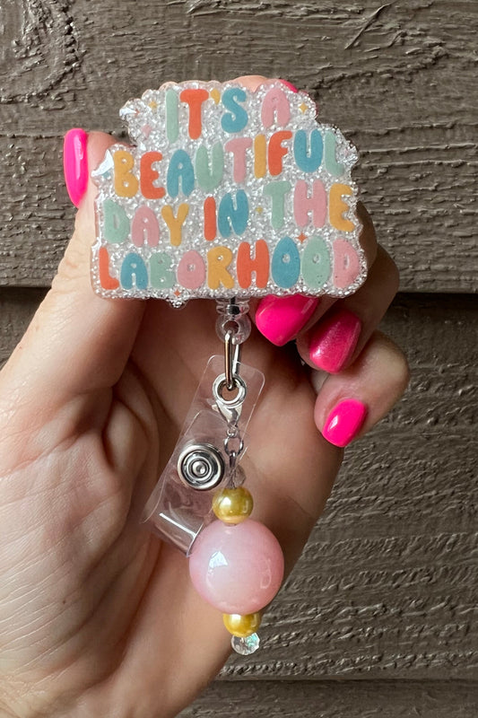 Soft Glitter Badge Reel featuring the phrase "It's a beautiful day in the laborhood" with soft pink and gold beaded dangle.