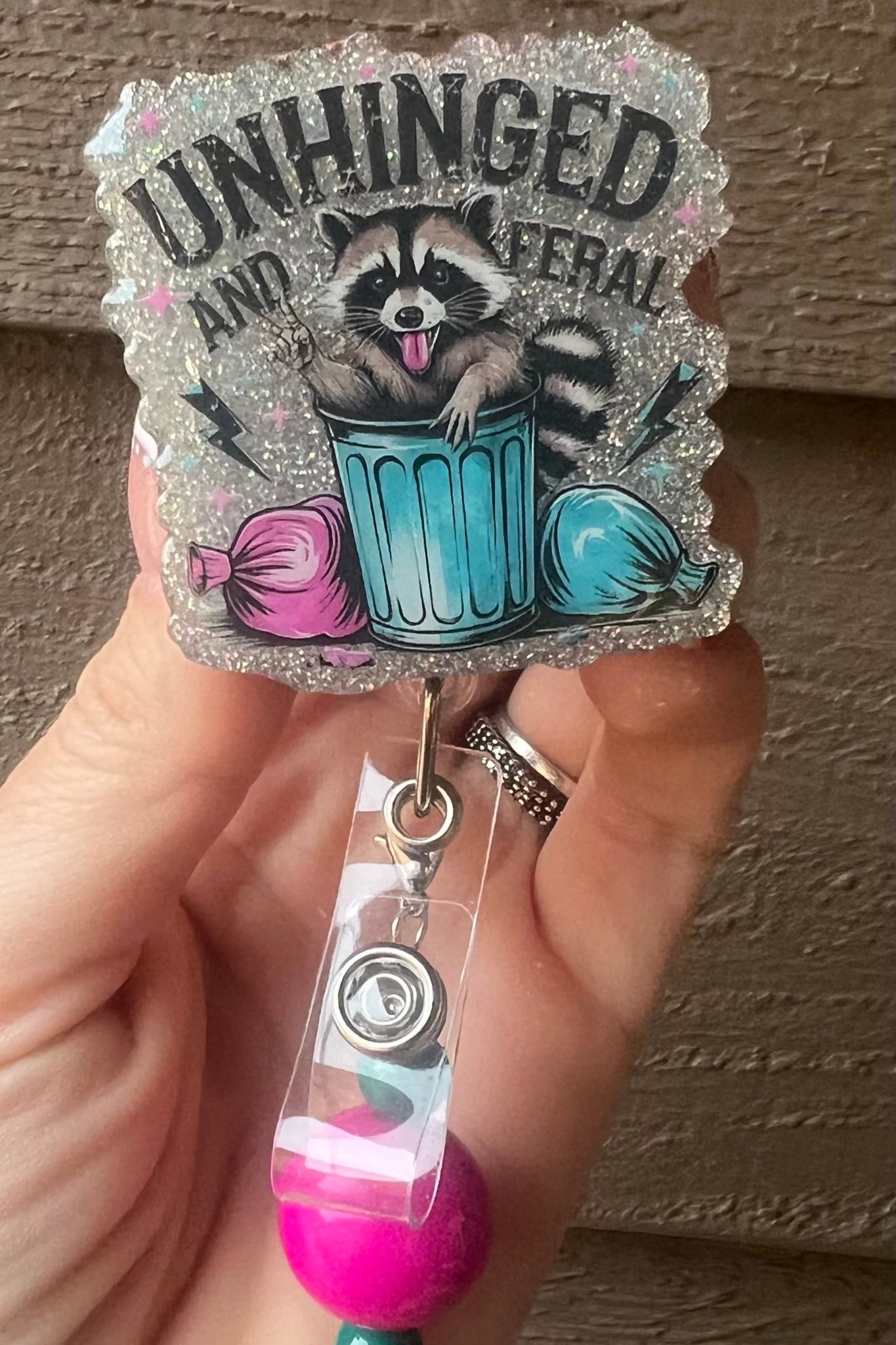 Glitter badge reel with a raccoon in a trash can flipping the bird, surrounded by garbage bags and the text ‘Unhinged and Feral
