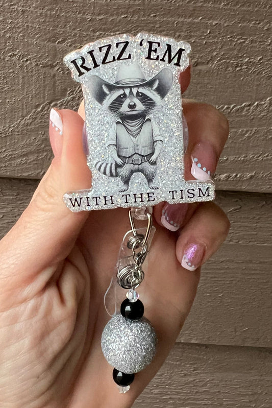 Funny Raccoon Badge Reel with "Rizz 'Em with the Tism" Printed over soft blue glitter.