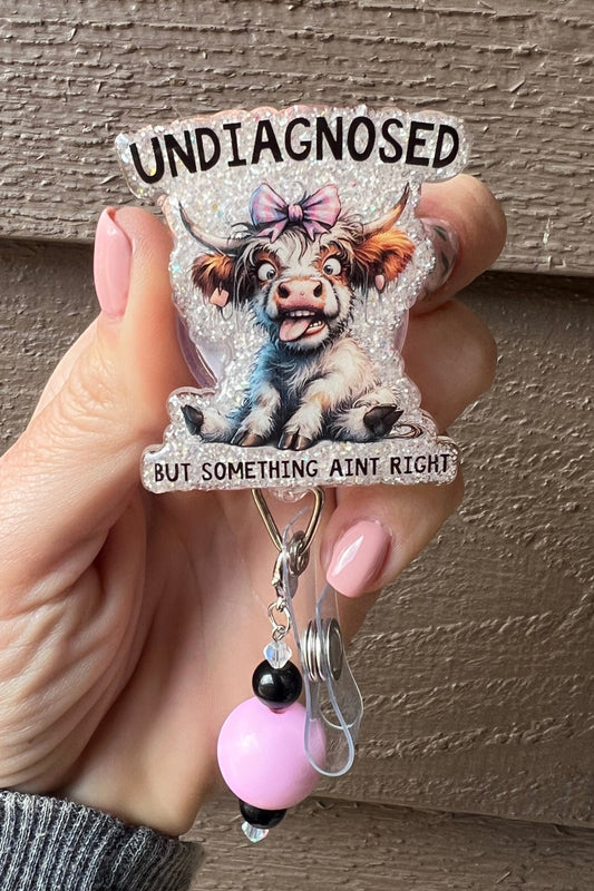 Funny Cow badge reel with pink bow and glitter background that says "Undiagnosed but something ain't right," with pink and black beaded charm.