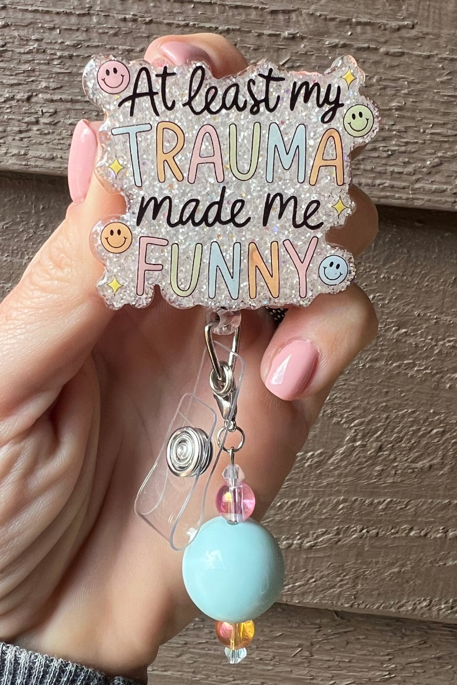 Pastel badge reel with soft glitter background and beaded charm that reads "At least my trauma made me funny," decorates with smiley faces and sparkle accents.
