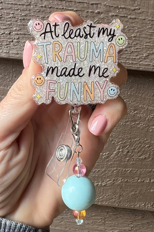 Pastel badge reel with soft glitter background and beaded charm that reads "At least my trauma made me funny," decorates with smiley faces and sparkle accents.