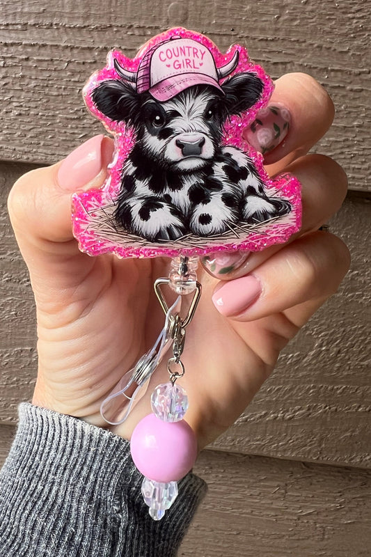 A pink glitter border badge reel with a cartoon cow wearing a pink "Country Girl" hat, decorated with a coordinating badge bling dangle.