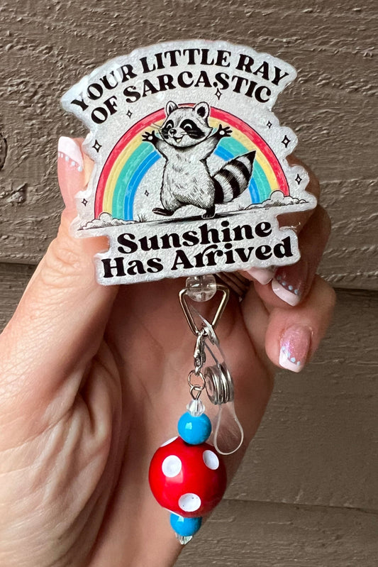 Rainbow Badge Reel with "Your Little Ray of Sarcastic Sunshine has Arrived" with a cheerful raccoon.