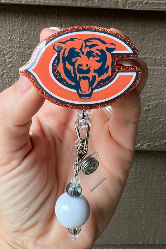 Chicago bears badge reel bordered with orange glitter and a white and navy beaded dangle