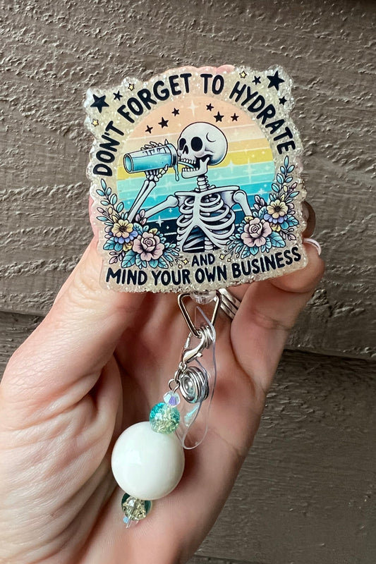 A soft yellow glittery badge reel with a rainbow background and skeleton drinking water, surrounded by flowers, with the text "Don't forget to hydrate and mind your own business," accented with coordinating badge bling.