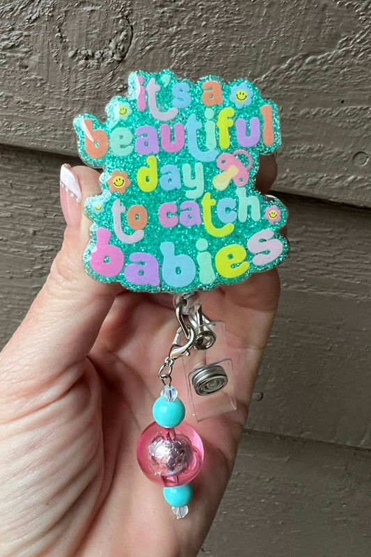 A Seafoam Glitter Background badge reel with the hrase "It's a beautiful day to catch babies" in colorful bubble letters, decorated with smiley faces and a coordinating badge bling.