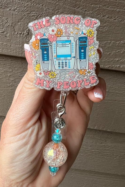 A glitter badge reel featuring IV pumps and the phrase "The Song of My People" surrounded by flowers and music notes, with coordinating badge bling.