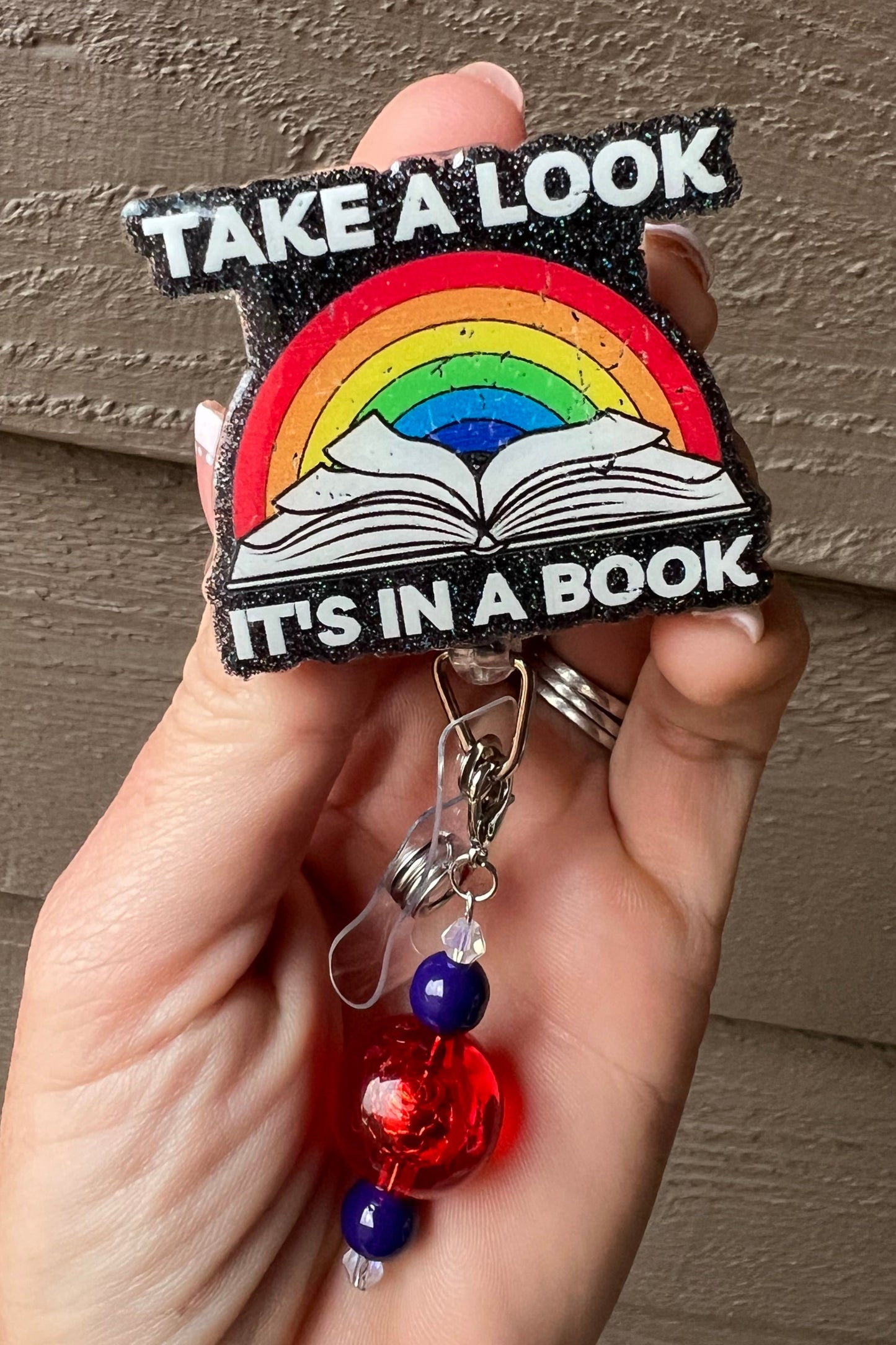 Holographic Black Glitter Background with a rainbow over an open book and the phrase "Take a Look, It's in a Book," accented with coordinating badge bling.