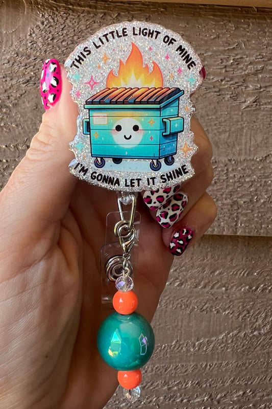 Smiling Dumpster on Fire Badge Reel with "This little light of mine i'm gonna let it shine" printed over soft blue glitter.