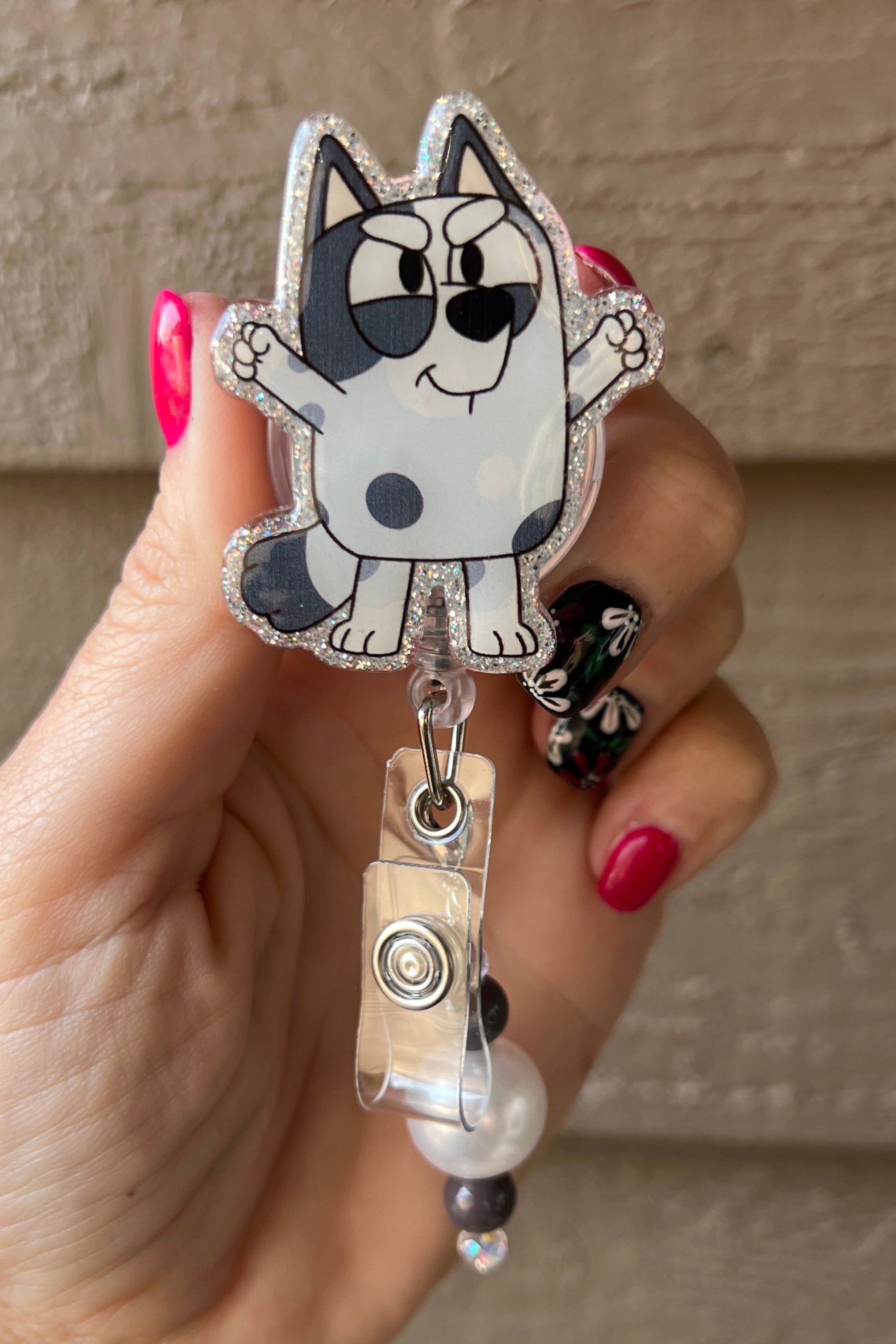 Glittery badge reel featuriing Muffin from Bluey with black, white, and pearl beaded dangle