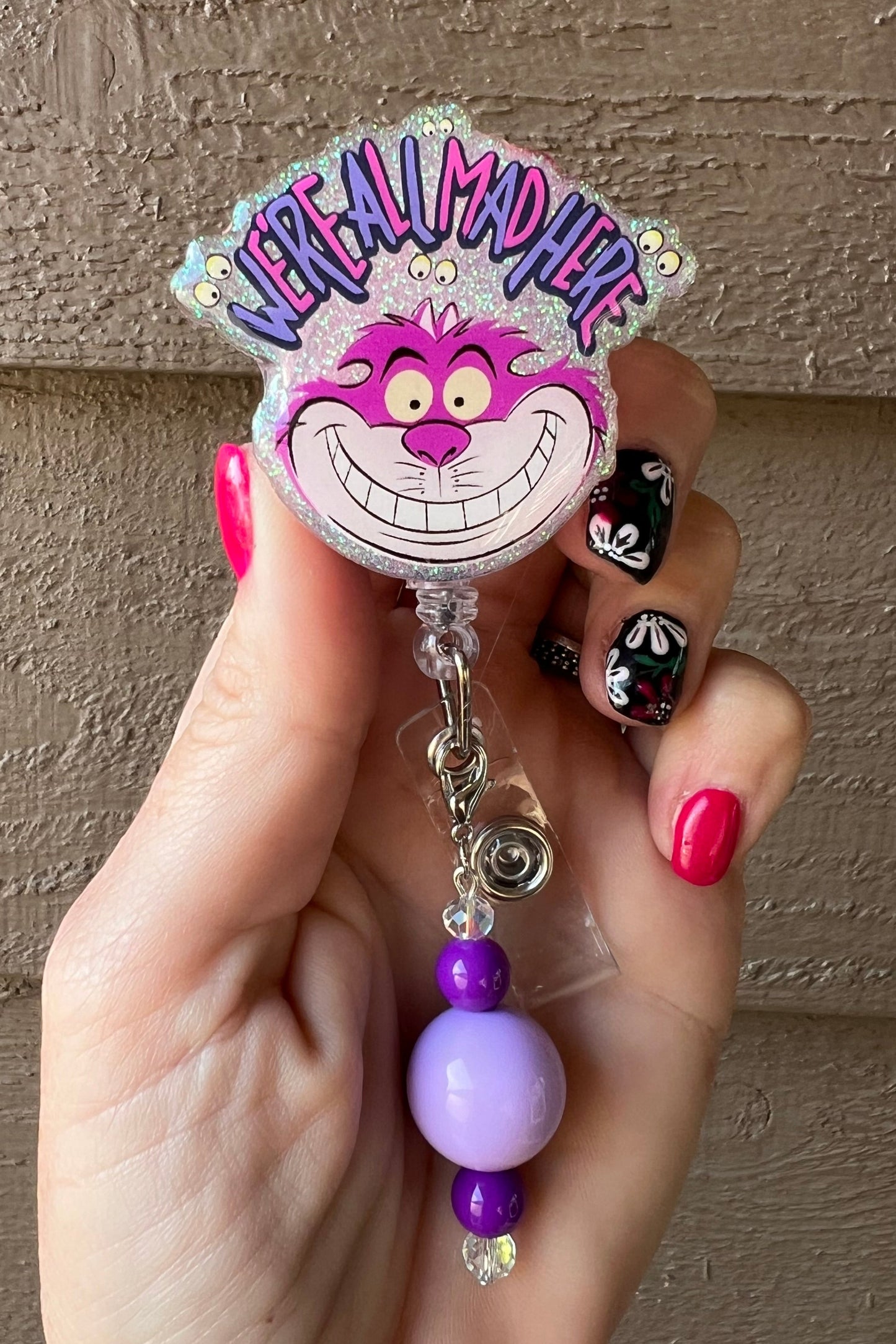 Glittery badge reel with Cheshire Cat smiling under "We're All Mad Here" text, purple beaded dangle
