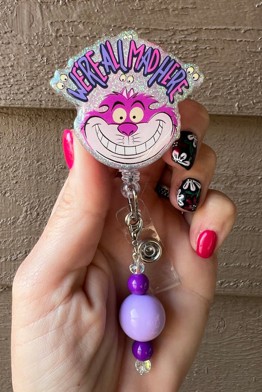 Glittery badge reel with Cheshire Cat smiling under "We're All Mad Here" text, purple beaded dangle