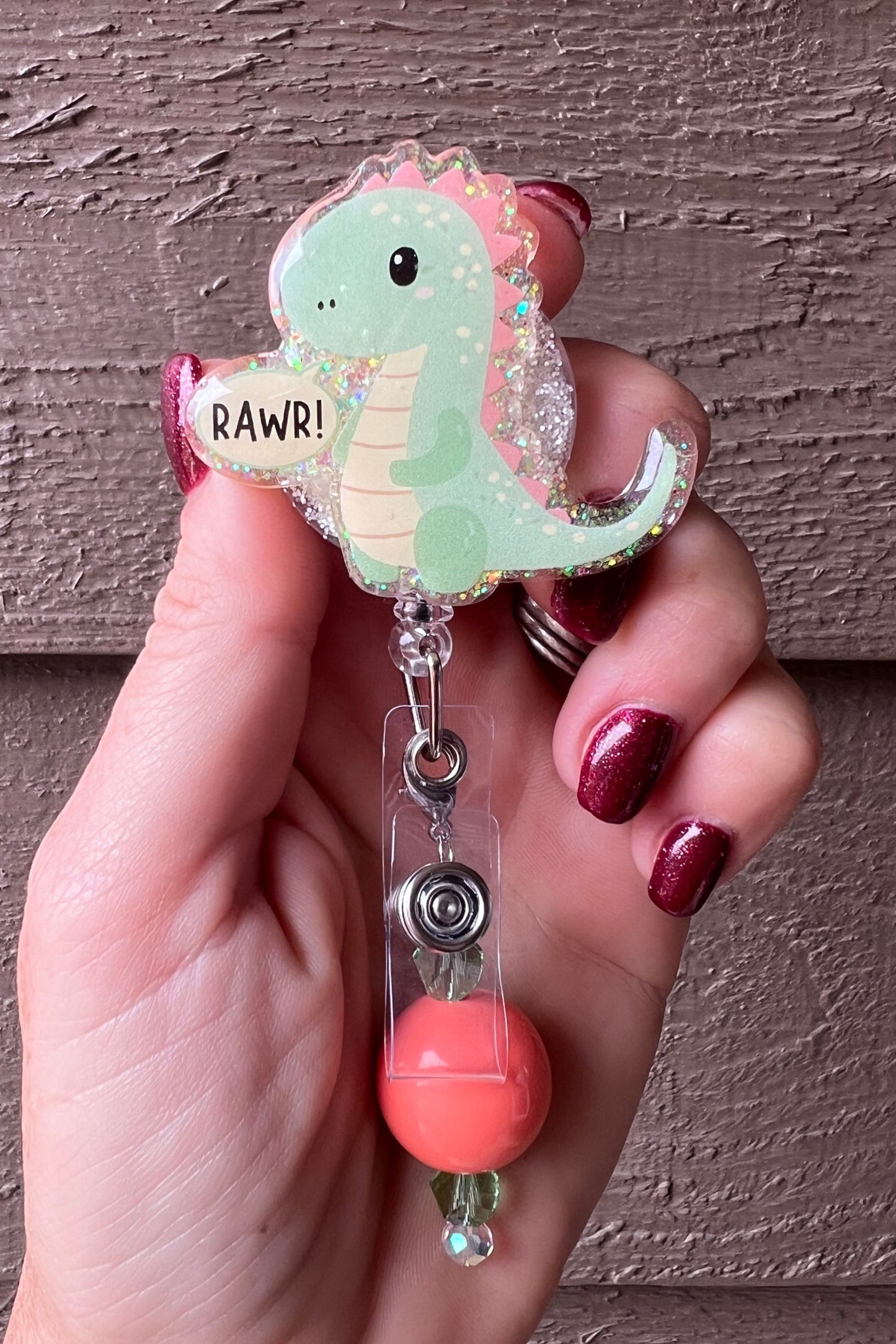 Hand holding a glittery badge reel featuring a pastel green cartoon dinosaur with pink spikes and a speech bubble that says “RAWR!”.