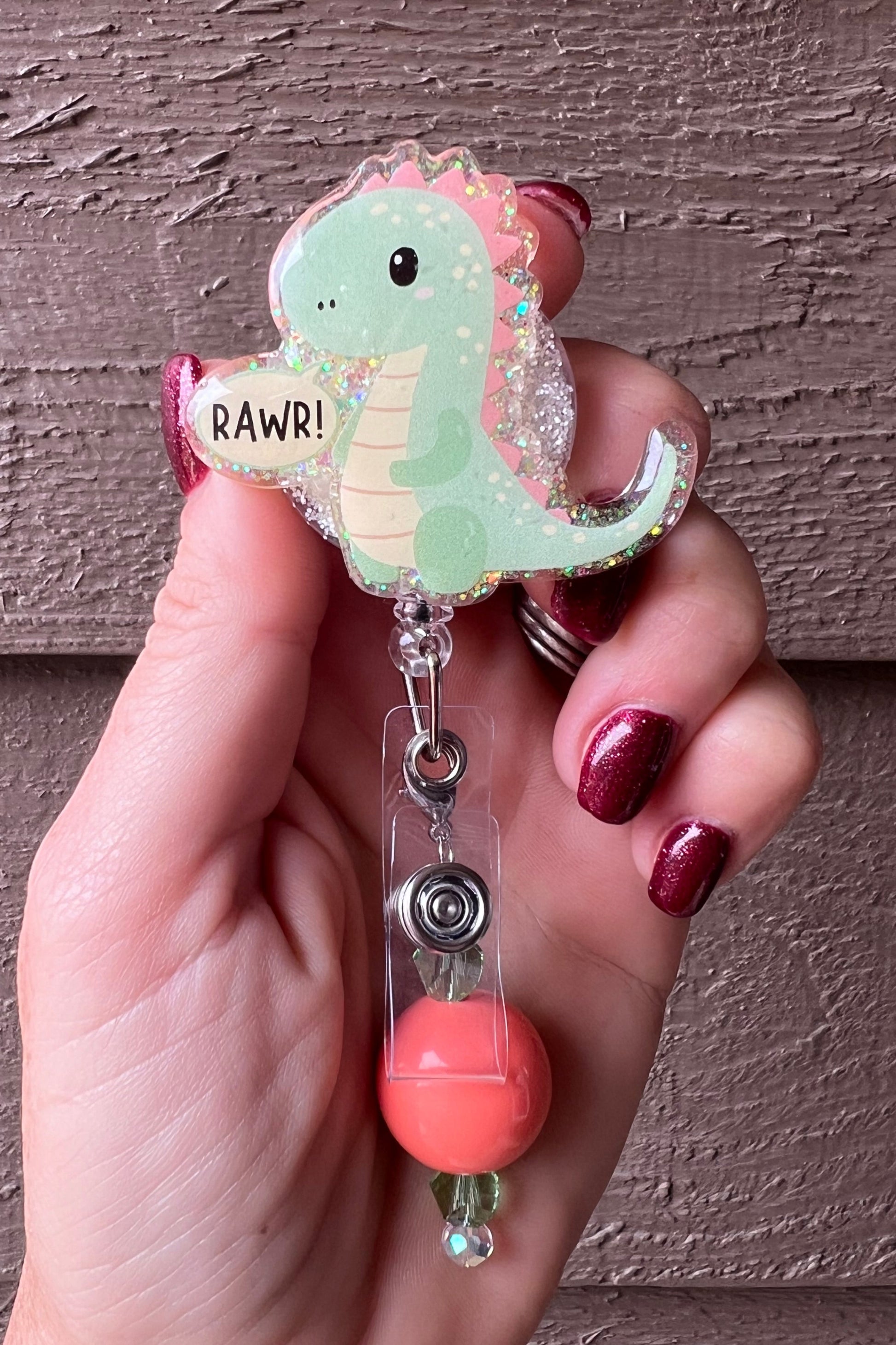 Hand holding a glittery badge reel featuring a pastel green cartoon dinosaur with pink spikes and a speech bubble that says “RAWR!”.