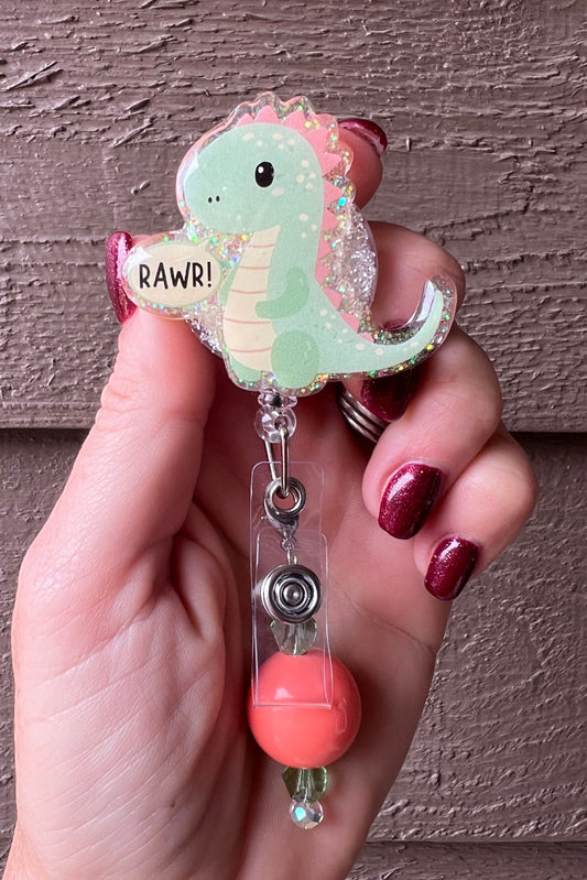 Hand holding a glittery badge reel featuring a pastel green cartoon dinosaur with pink spikes and a speech bubble that says “RAWR!”.