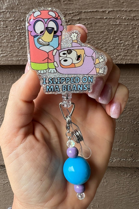 Badge reel featuring Bluey and Bingo as the Grannies with "I Slipped on a Beans" in text on glitter background with blue and purple dangle.