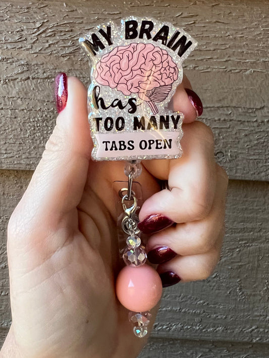 Hand holding a glitter badge reel with a pink brain design and the phrase “My Brain Has Too Many Tabs Open,” featuring a dangle with peach and clear crystal beads, against a textured wooden background.