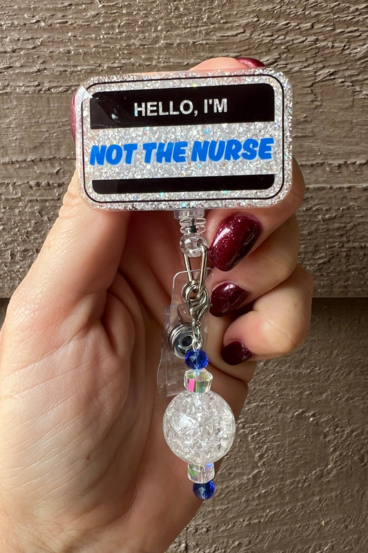 Hand holding a glittery rectangular badge reel with the text “Hello, I’m NOT THE NURSE” in bold black and blue lettering.