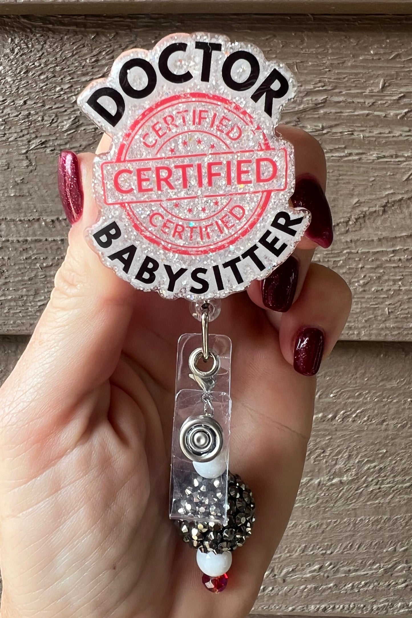 Hand holding a glittery badge reel with bold black text reading “Doctor Babysitter” and a large red stamp that says “Certified"