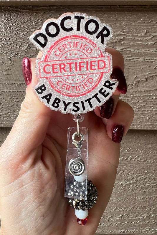 Hand holding a glittery badge reel with bold black text reading “Doctor Babysitter” and a large red stamp that says “Certified"