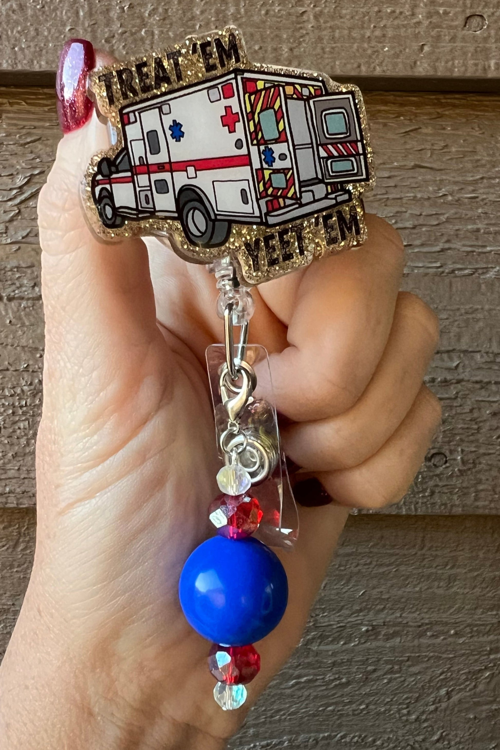 Hand holding a glittery badge reel featuring a cartoon ambulance and the phrase “Treat ’Em Yeet ’Em,” with a dangle of red, blue, and clear beads, set against a wood-textured background.