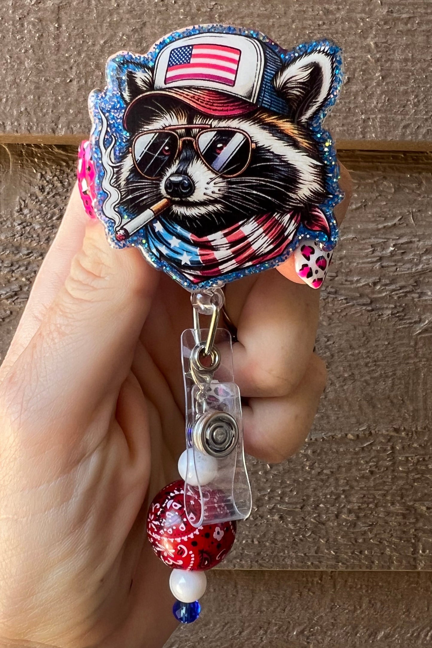 Patriotic raccoon badge reel with American flag hat and bandana, wearing sunglasses and holding a cigar. Beaded charm with a red bandana bead, white and blue accents. Held in hand with pink leopard print nails against a wood background.