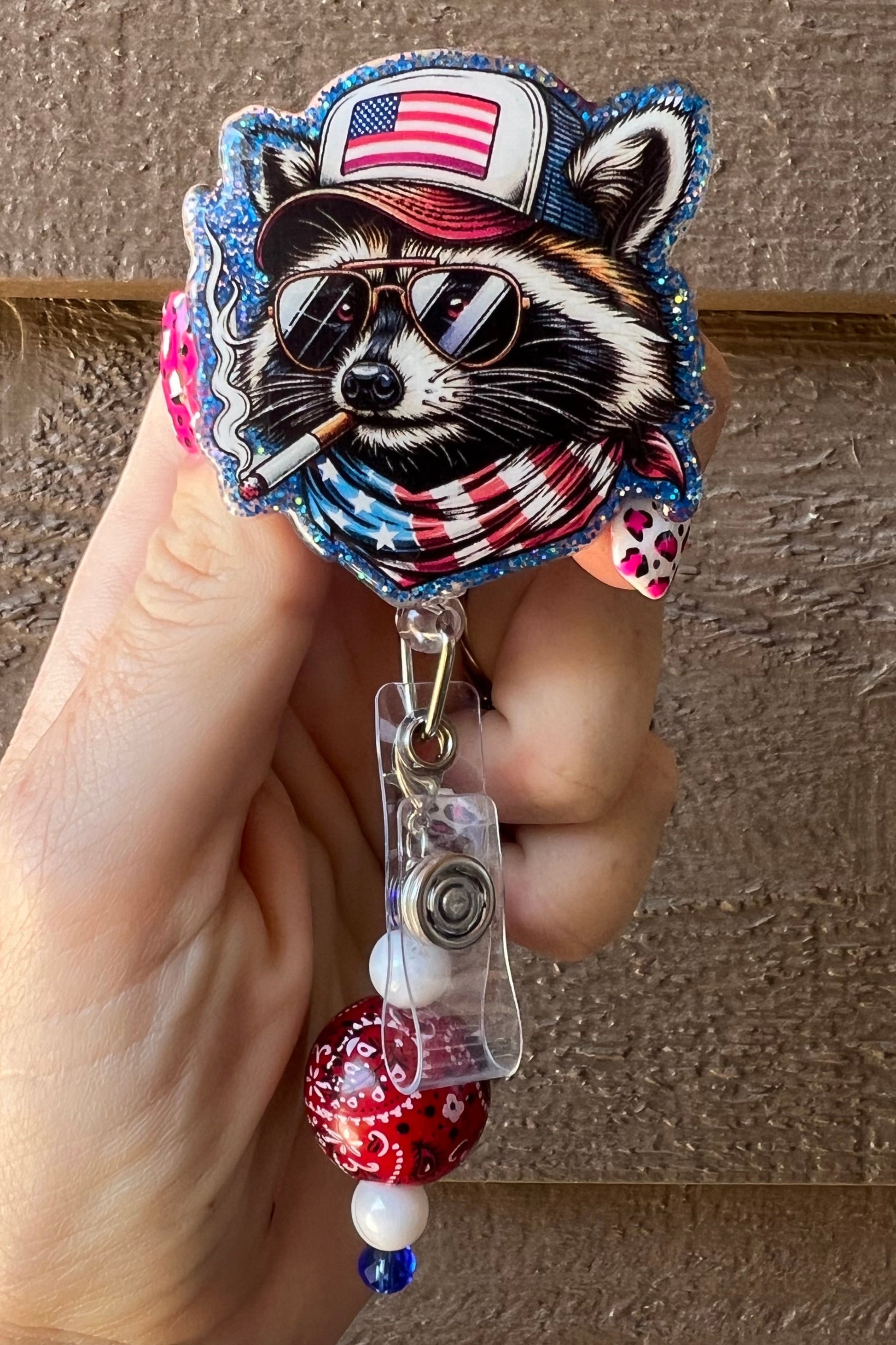 Patriotic raccoon badge reel with American flag hat and bandana, wearing sunglasses and holding a cigar. Beaded charm with a red bandana bead, white and blue accents. Held in hand with pink leopard print nails against a wood background.