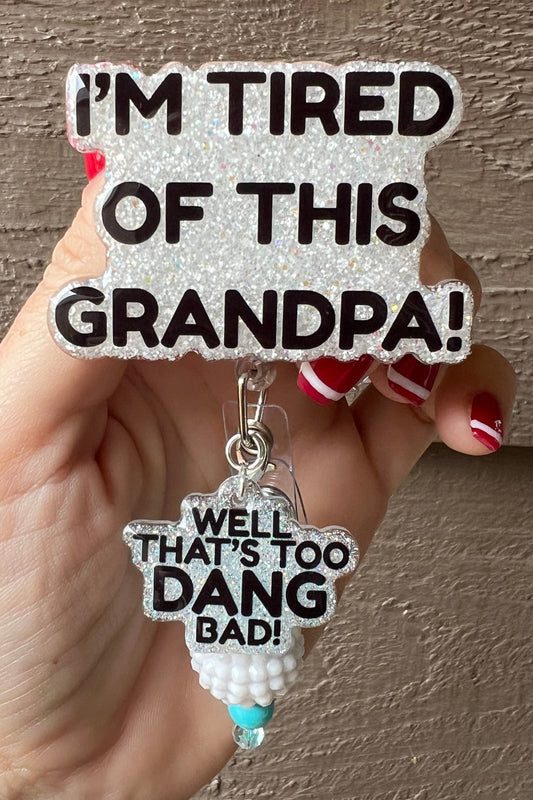 Hand holding a glittery badge reel with the quote “I’m Tired of This Grandpa!” and a hanging charm that says “Well That’s Too Dang Bad!” with beaded accent