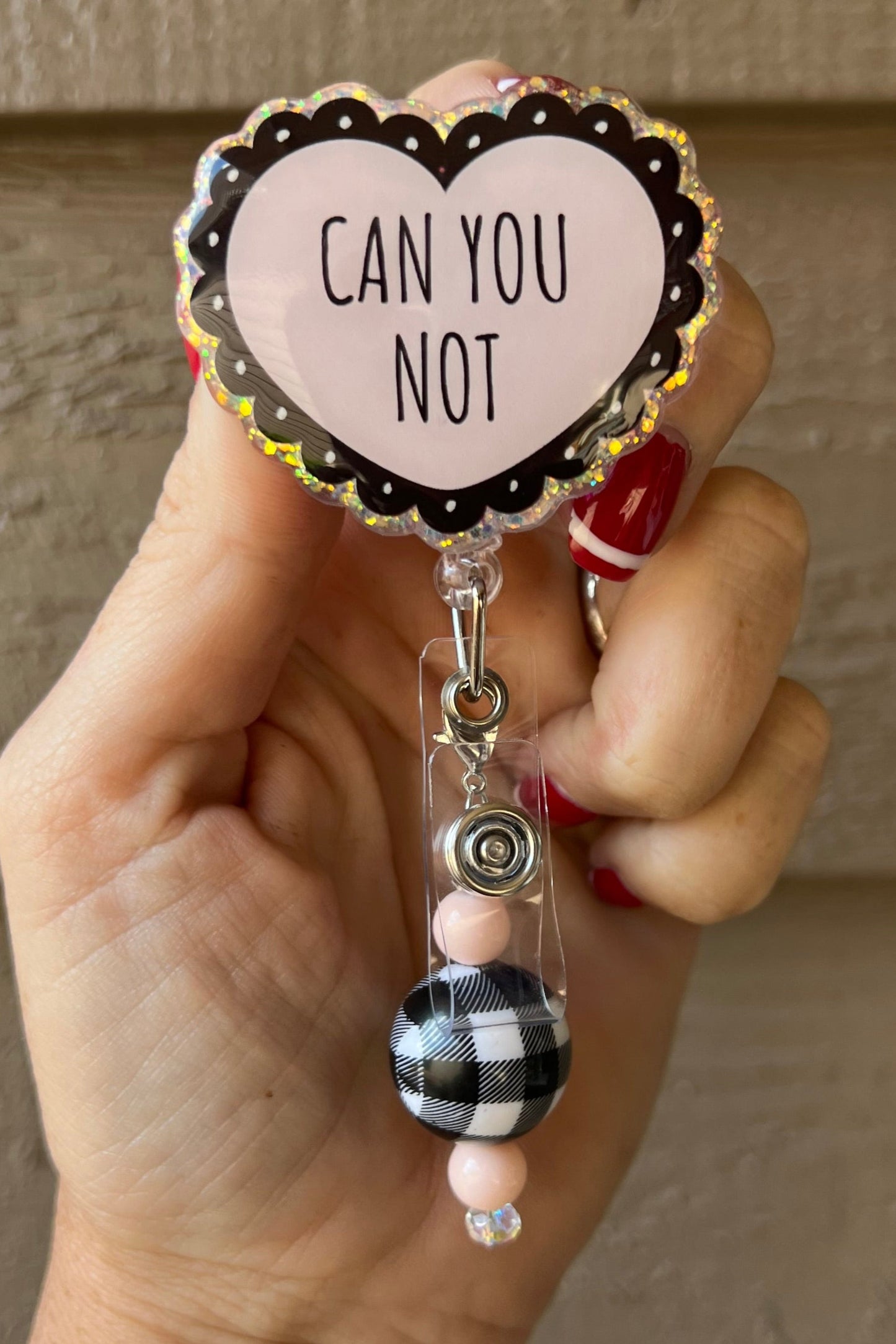 Hand holding a holographic heart-shaped badge reel with “Can You Not” text and plaid/pink beaded charm