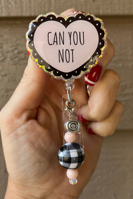 Hand holding a holographic heart-shaped badge reel with “Can You Not” text and plaid/pink beaded charm