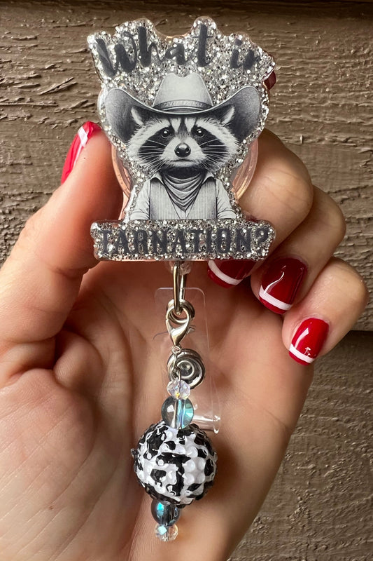 Hand holding a glittery badge reel with a raccoon in a cowboy hat and “What in Tarnation” text, decorated with a black-and-white cow print bead charm.