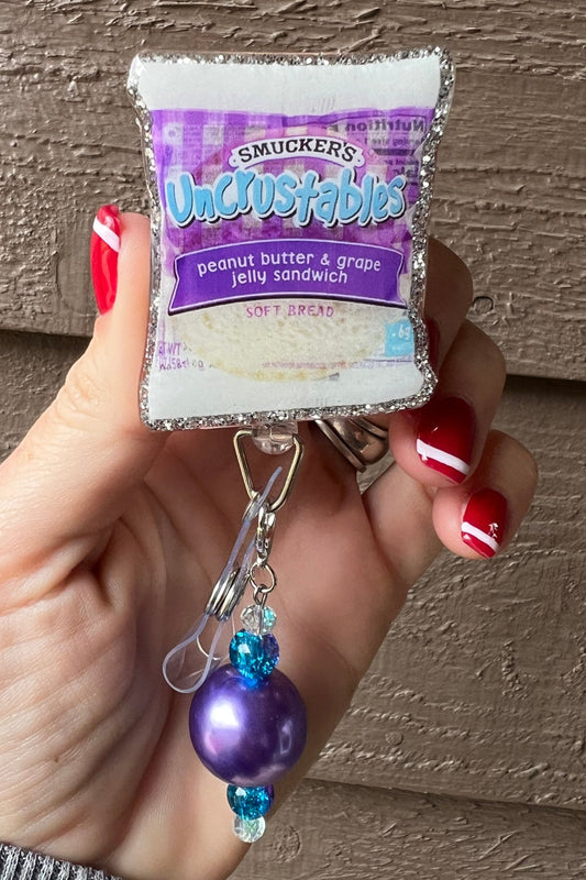 uncrustables badge reel with glittery PB&J design and purple beaded dangle.