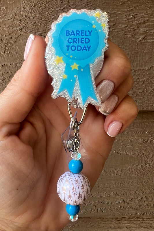 Hand holding a glittery blue badge reel shaped like a ribbon that says “Barely Cried Today,” with blue and white beaded charm attached, set against a wooden background.
