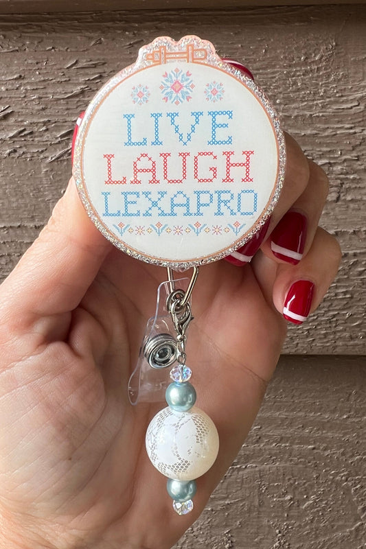 Hand holding a glittery badge reel featuring a cross-stitch style design reading “Live Laugh Lexapro” with teal and white beaded charm