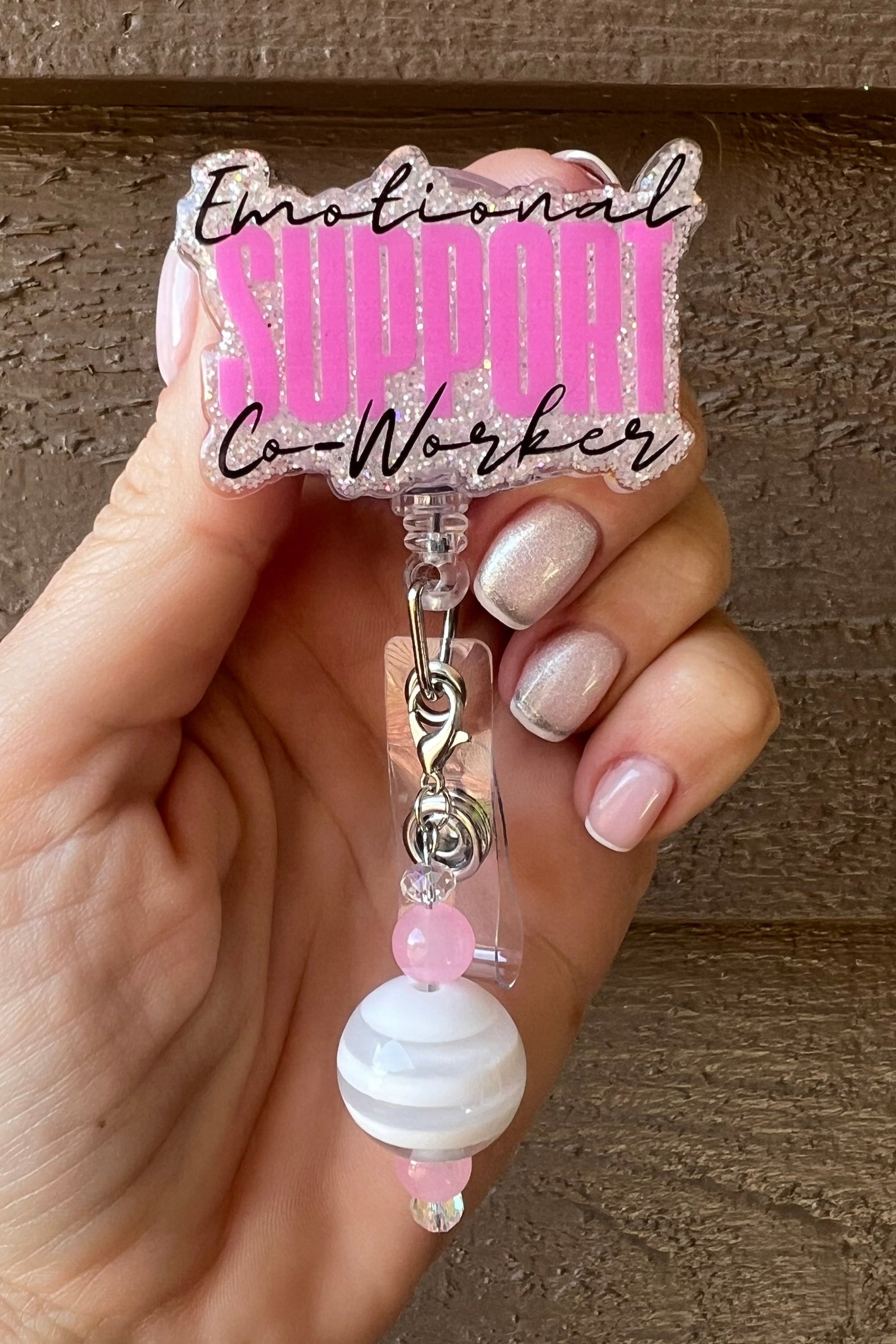 Hand holding a pink glitter badge reel with the words “Emotional Support Co-Worker” in black and bold pink lettering. The reel features a beaded charm with white and pink beads, clipped to a retractable badge holder, shown against a wood background.
