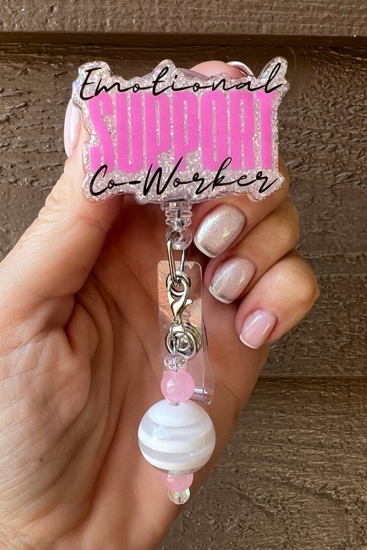 Hand holding a pink glitter badge reel with the words “Emotional Support Co-Worker” in black and bold pink lettering. The reel features a beaded charm with white and pink beads, clipped to a retractable badge holder, shown against a wood background.
