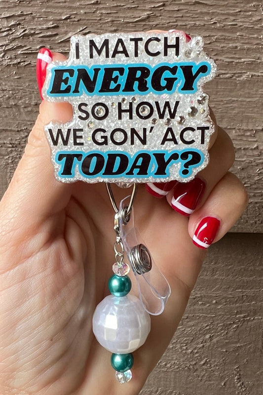 Hand holding badge reel that says "I match energy so how we gon' act today" on pearl white glitter border with rhinestones and matching badge bling.